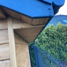 Fox House - Fragment nowej, drewnianej konstrukcji z grafitowym pokryciem dachowym i rynną, widoczny z dołu, na tle ogrodzenia i zieleni.