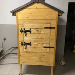 Drewniana wędzarnia z dachem i termometrem, wykonana w Myślenicach, stoi na kółkach w pomieszczeniu gospodarczym.