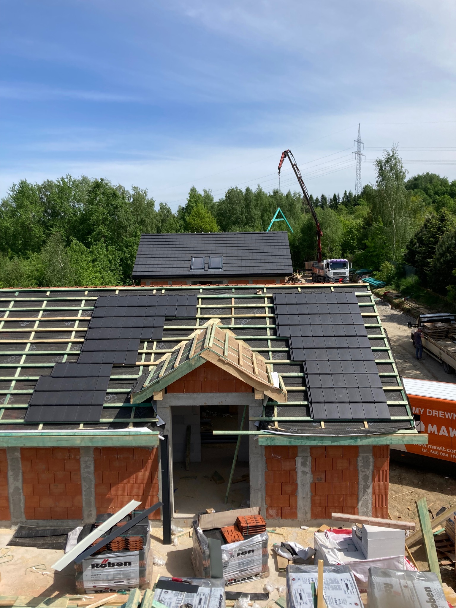 Montaż czarnej dachówki ceramicznej na nowym budynku, widoczna konstrukcja więźby dachowej i częściowo ułożone pokrycie, w tle dźwig budowlany i zieleń drzew.