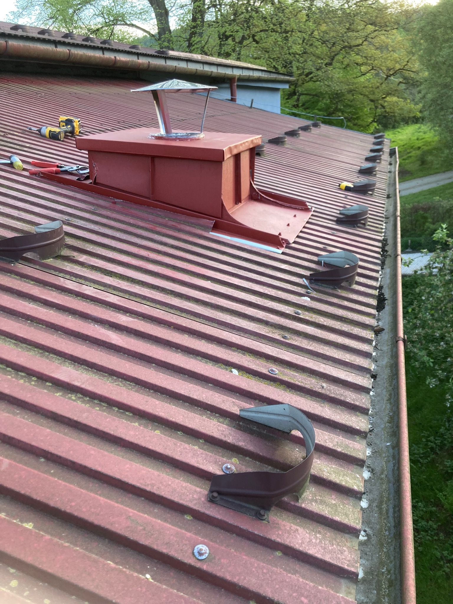 Czerwony blaszany dach z kominem, wentylatorami i narzędziami dekarza, widok z góry na tle zieleni.