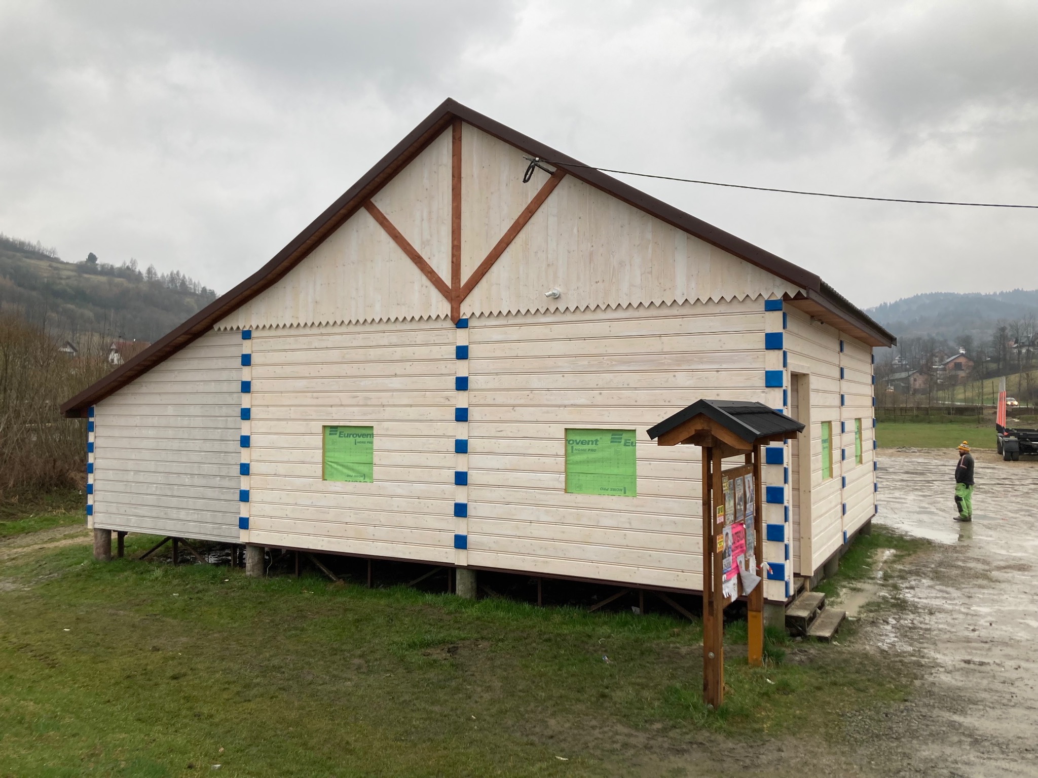 Nowa drewniana konstrukcja budynku z widoczną więźbą dachową, obita jasnymi deskami z niebieskimi akcentami, na podwyższeniu, z widocznymi oknami zabezpieczonymi folią i słupem ogłoszeniowym obok...