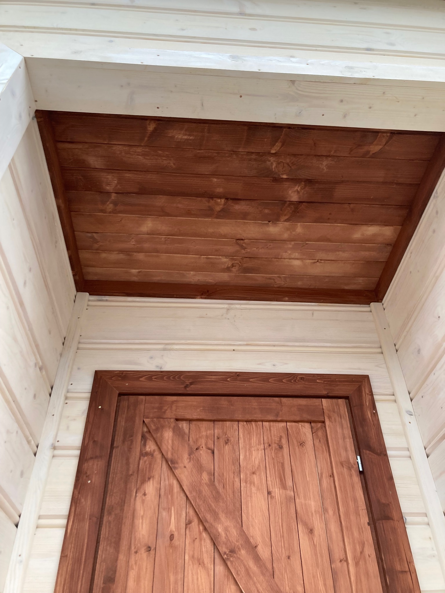 Drewniana konstrukcja altany z brązowym, lakierowanym sufitem i drzwiami typu stodoła, widoczne elementy z jasnego, surowego drewna.