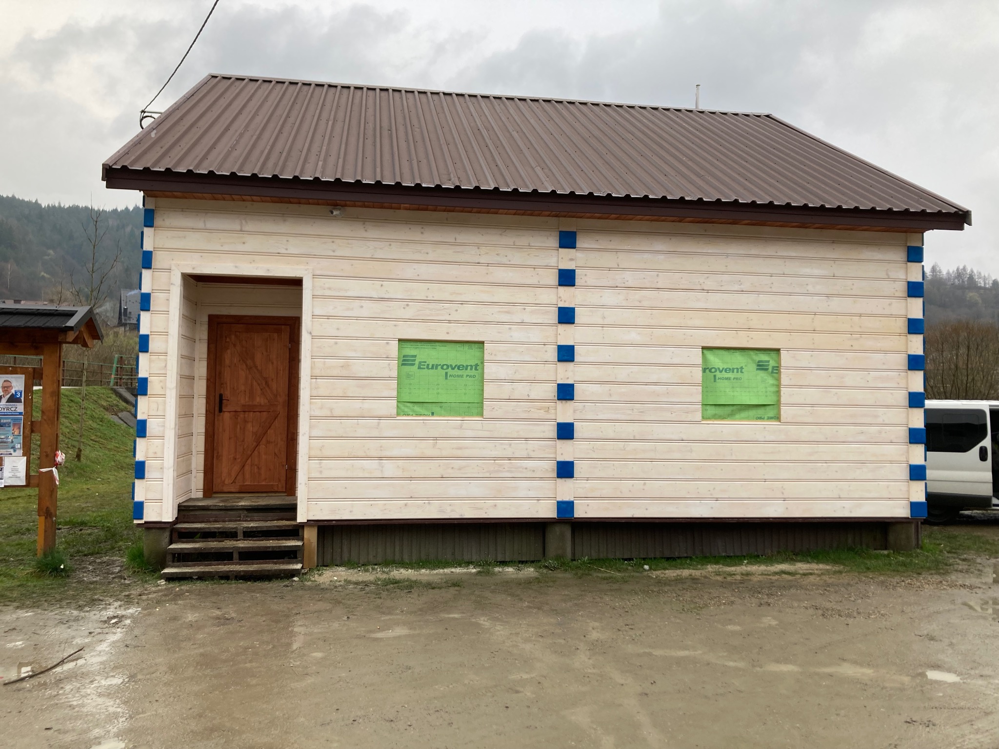 Nowo wybudowany drewniany budynek z brązowym dachem z blachy falistej, zabezpieczone okna folią Eurovent, widoczne góry w tle.