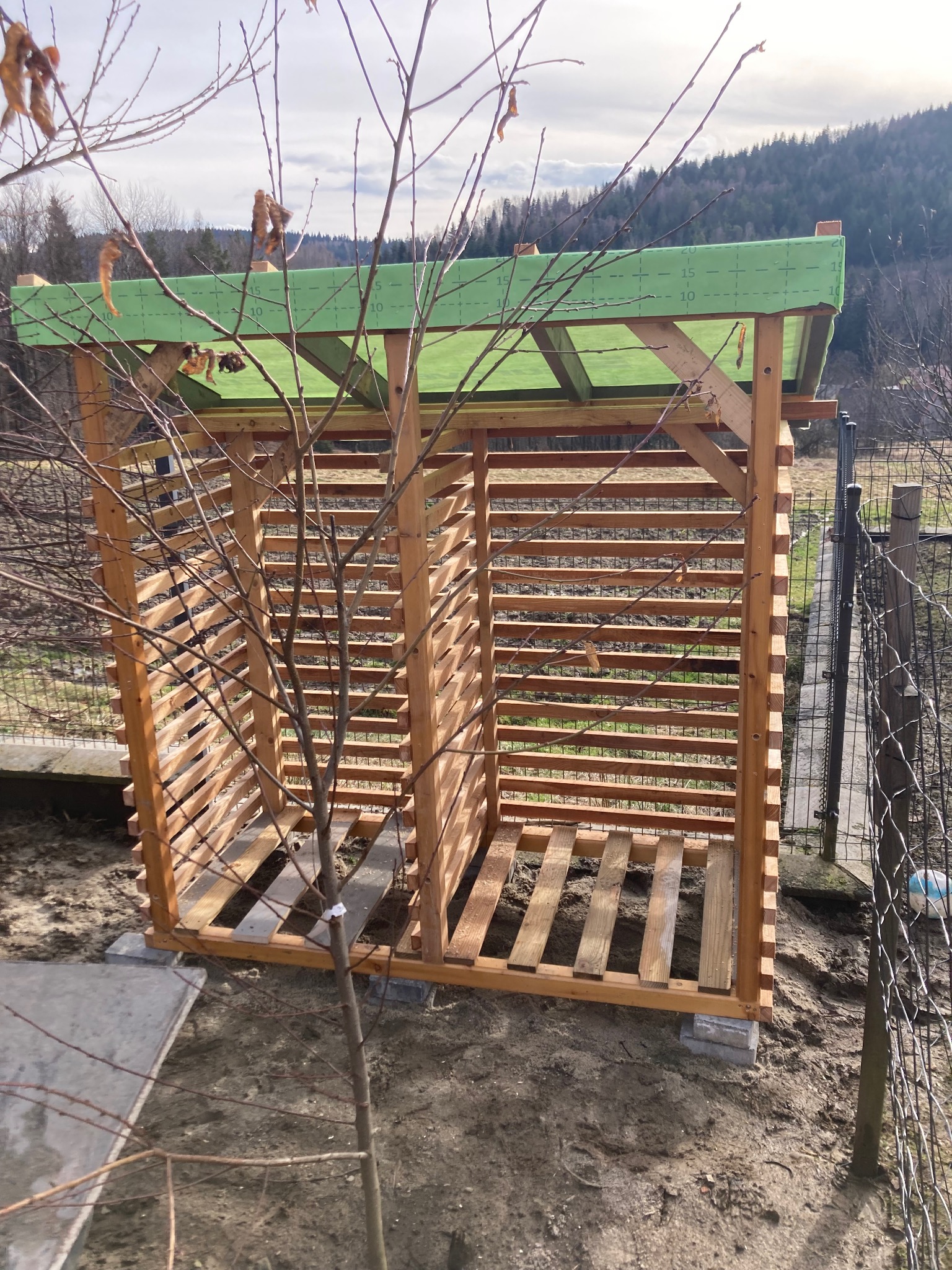 Nowa, drewniana konstrukcja altany z zielonym, częściowo przezroczystym dachem, w surowym stanie, na tle górskiego krajobrazu i młodego drzewa bez liści.