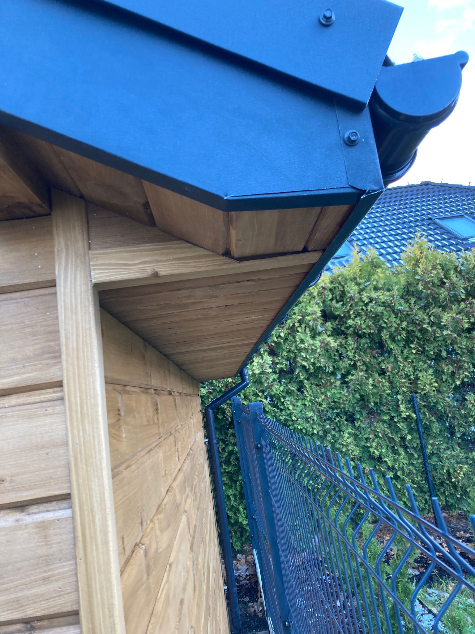 Fragment nowej, drewnianej konstrukcji z grafitowym pokryciem dachowym i rynną, widoczny z dołu, na tle ogrodzenia i zieleni.