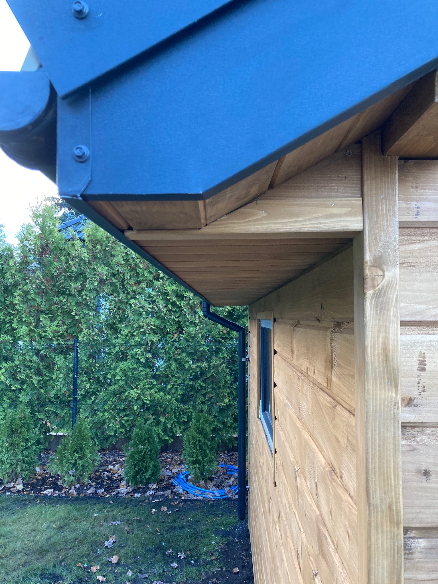 Detale konstrukcyjne drewnianej altany z ciemnym, metalowym zadaszeniem i rynną, widok z boku na tle zielonych tuj.