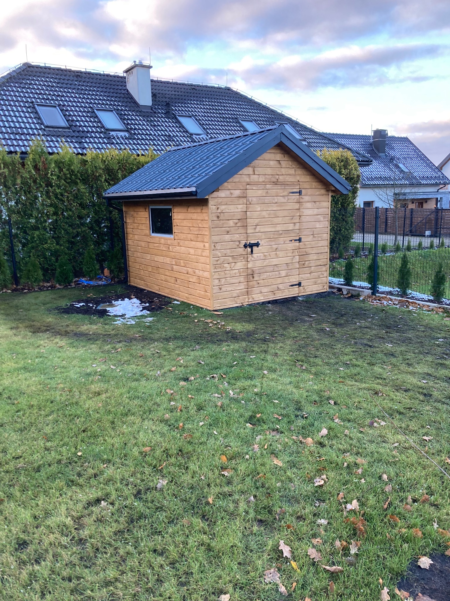Nowa, drewniana altana ogrodowa z szarym dachem, umieszczona na trawniku z resztkami śniegu, z widocznym domem w tle.