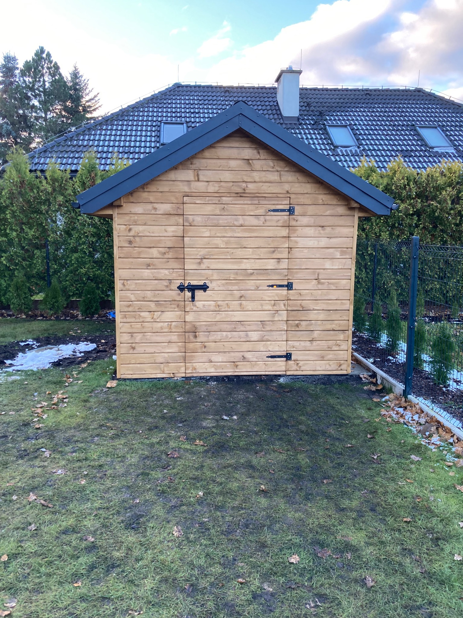 Drewniana altana ogrodowa z czarnymi okuciami na tle domu z ciemną dachówką i zielonymi tujami.