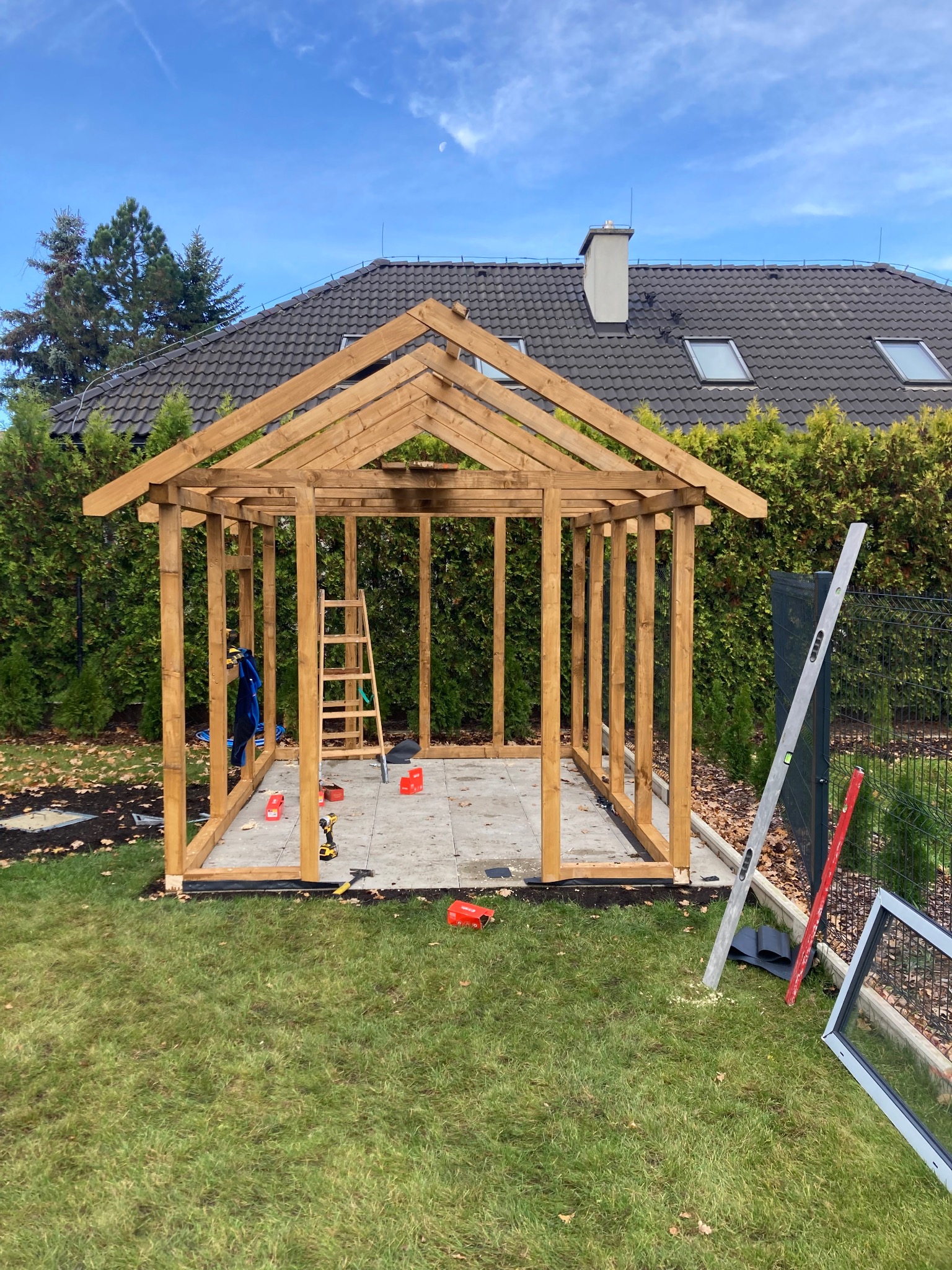 Konstrukcja drewnianej altany w trakcie budowy, widoczne narzędzia, drabina oparta o konstrukcję, poziomica oparta o ogrodzenie, trawa, krzewy, dom z ciemnym dachem w tle.
