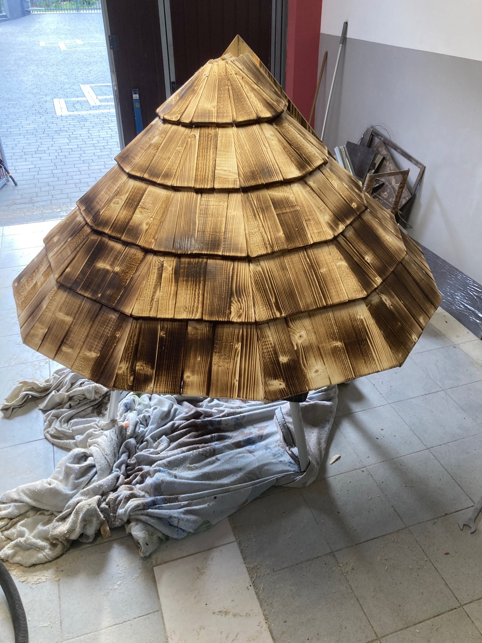 Drewniana konstrukcja w kształcie stożka, pokryta gontem, prawdopodobnie element altany, na tle garażu lub warsztatu.