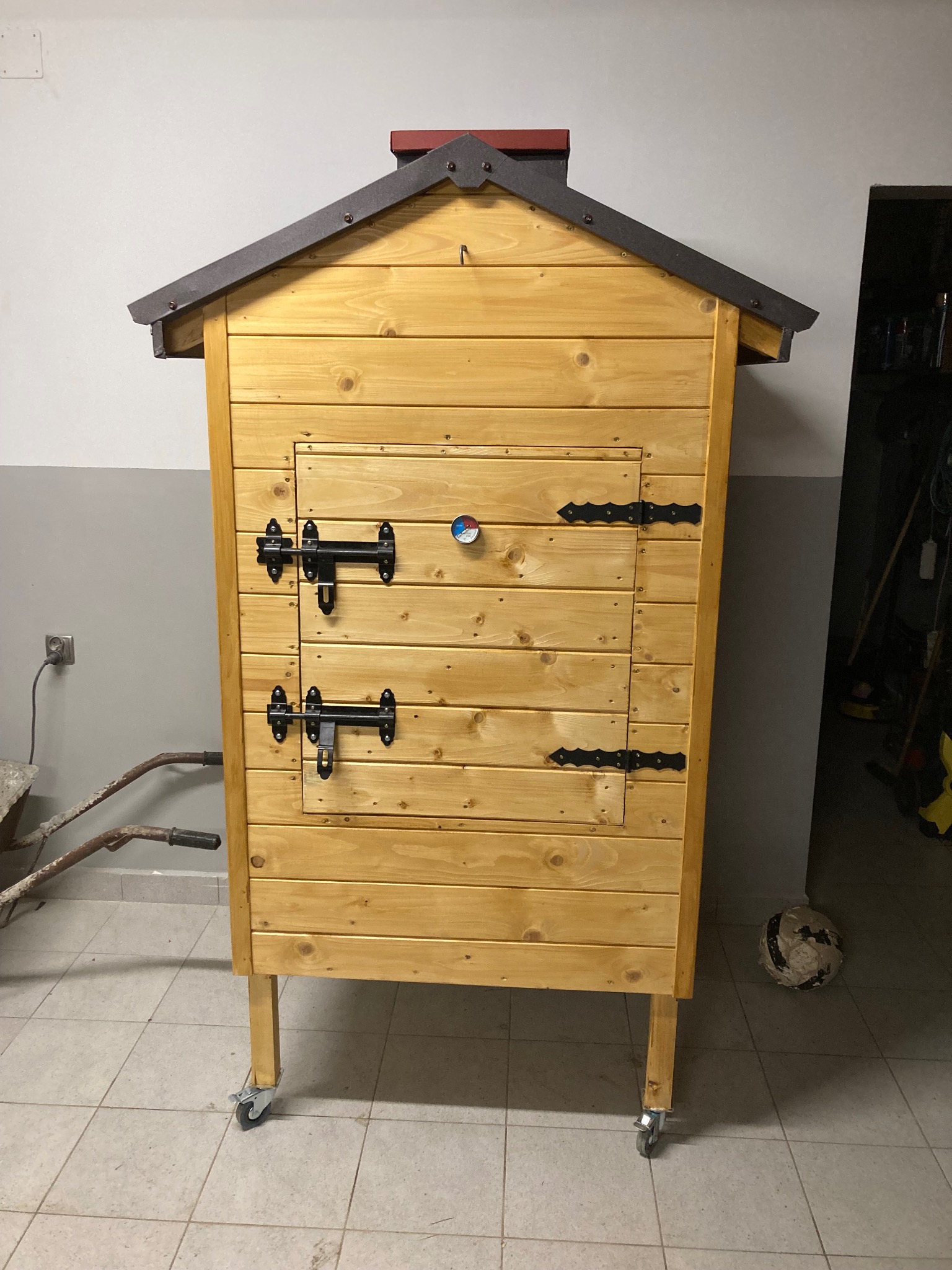 Drewniana wędzarnia z dachem i termometrem, wykonana w Myślenicach, stoi na kółkach w pomieszczeniu gospodarczym.