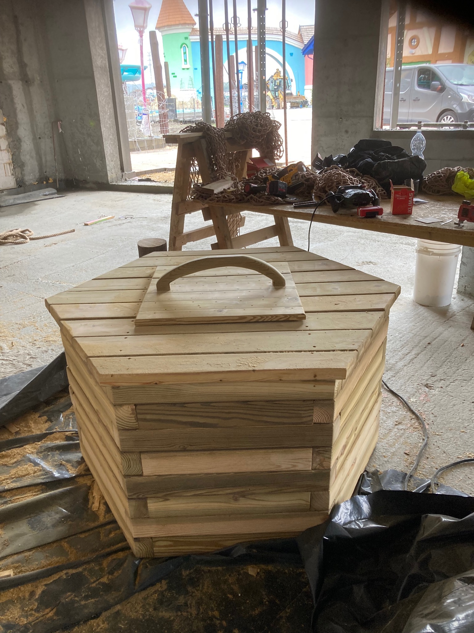 Nowa, surowa, drewniana konstrukcja w kształcie sześcioboku z uchwytem na pokrywie, otoczona folią ochronną i narzędziami w tle, w trakcie budowy placu zabaw.