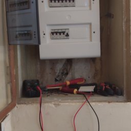 MORAS TOMASZ MORAWSKI - Otwarta skrzynka elektryczna z bezpiecznikami, multimetr, śrubokręty i rękawice robocze w zagłębieniu ściany podczas pomiarów elektrycznych.