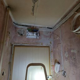 MORAS TOMASZ MORAWSKI - Rozdzielnia elektryczna z przewodami na ścianie w trakcie remontu, widoczna drabina i instalacja domofonowa. Ściany w stanie surowym, przygotowane do malowania.