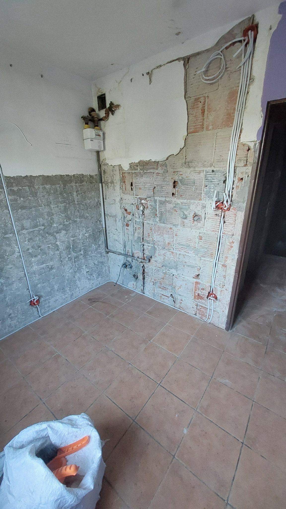 Wnętrze w trakcie remontu: odsłonięta cegła, nowe przewody elektryczne w peszlach, licznik gazu i narzędzia w worku na podłodze.
