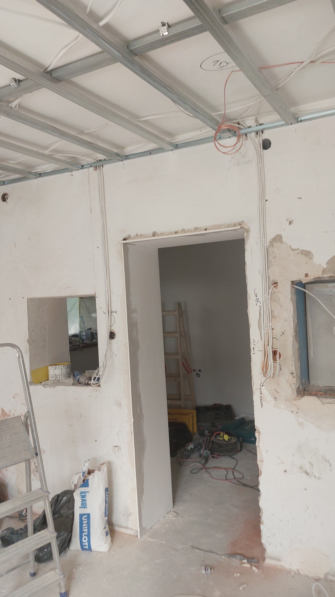 Wnętrze w trakcie remontu: sufit podwieszany na metalowej konstrukcji, wystające przewody elektryczne, ściany w stanie surowym, drabina i worki z zaprawą budowlaną w pomieszczeniu.