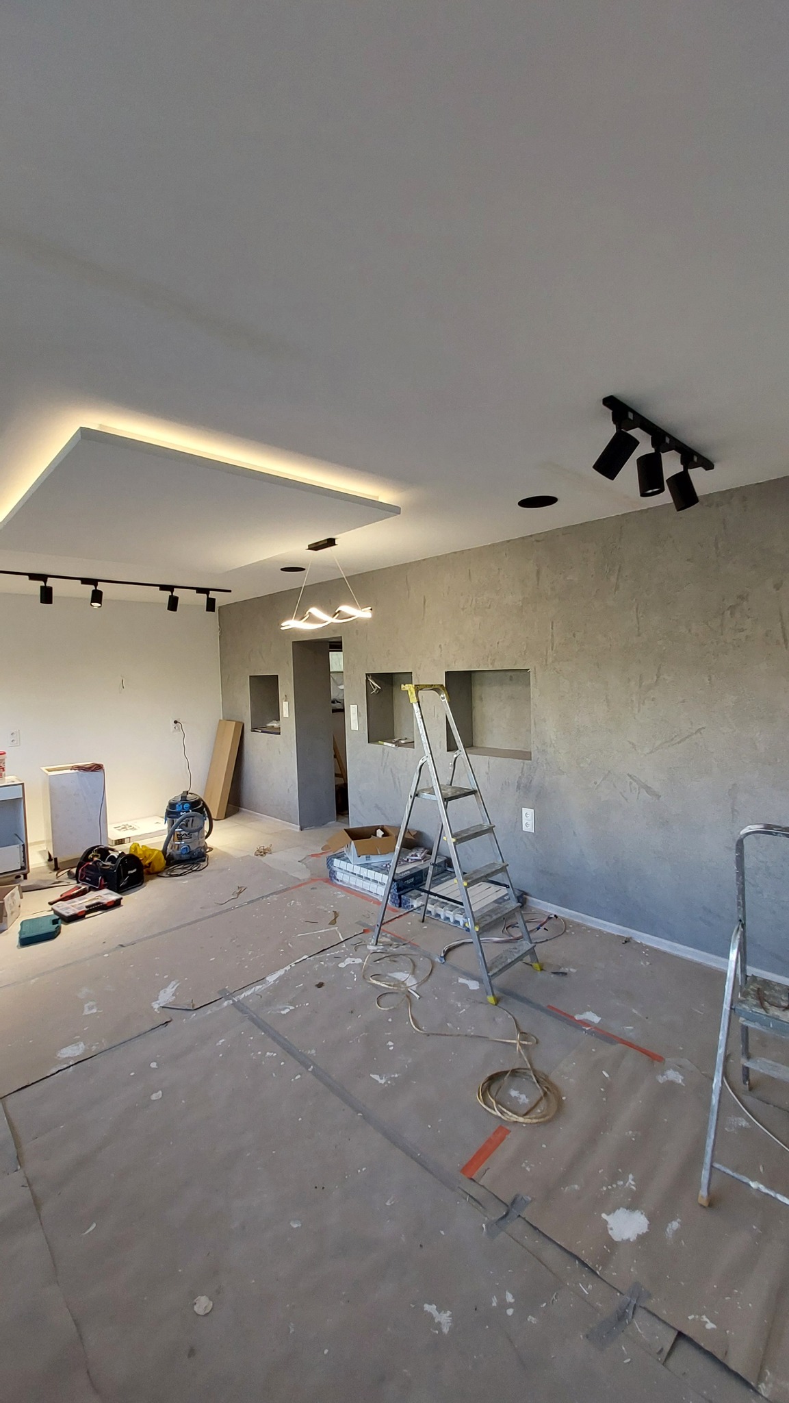 Wnętrze w trakcie remontu z widocznymi elementami oświetlenia sufitowego: listwa LED w podwieszanym suficie, reflektory szynowe oraz nowoczesna lampa wisząca. Na podłodze rozłożona folia ochronna...