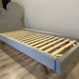 Szare ł&oacute;żko dziecięce z zagł&oacute;wkiem w kształcie chmury, z drewnianym stelażem, na drewnianej podłodze.