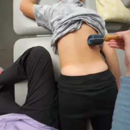 Pacjent leżący na brzuchu na stole rehabilitacyjnym, terapeuta używa rollera z kolcami na jego plecach, widoczne ubrania pacjenta i terapeuty.