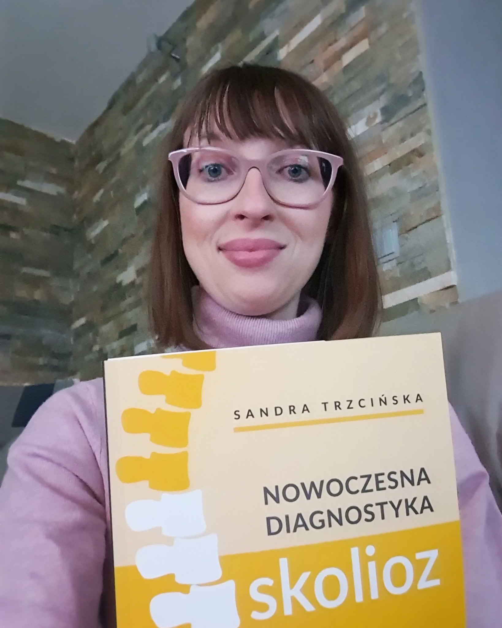 Uśmiechnięta kobieta w okularach trzyma książkę 'Nowoczesna Diagnostyka Skolioz' Sandry Trzcińskiej, na tle ściany z kamienną okładziną.