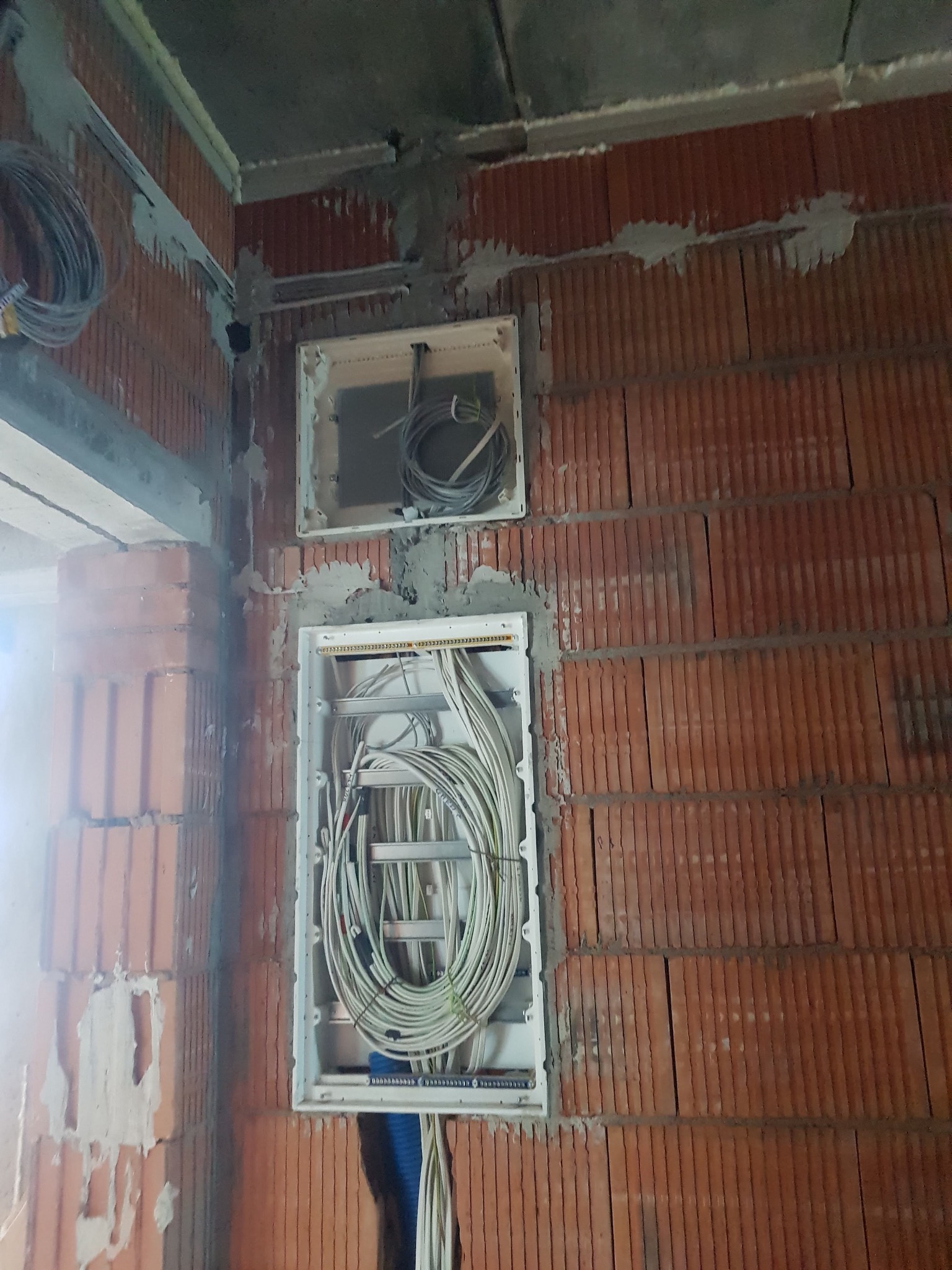 Wnętrze w trakcie instalacji elektrycznej: widoczne puszki instalacyjne w surowej, ceglanej ścianie, z poprowadzonymi kablami elektrycznymi, częściowo zwiniętymi wewnątrz puszek.