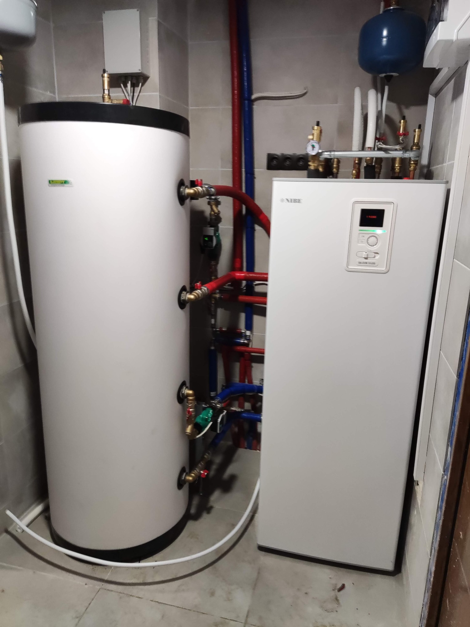 Biały zbiornik akumulacyjny i pompa ciepła Nibe z podłączonymi rurami w kotłowni w Wargowie, widoczne płytki ceramiczne na ścianach i podłodze.