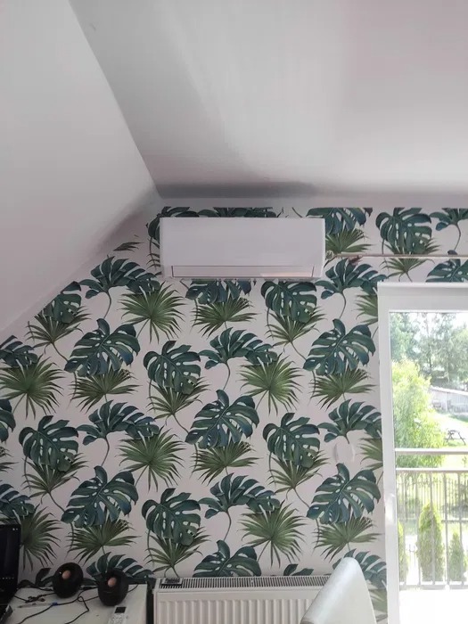 Biała jednostka klimatyzacyjna na ścianie z tapetą w tropikalne liście, widok na zewnątrz przez okno, pod skosem dachu.