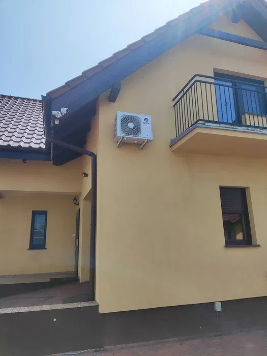 Żółta elewacja domu jednorodzinnego z zamontowanym klimatyzatorem marki Gree na ścianie, widoczny balkon z czarną balustradą i system kamer monitoringu pod dachem.