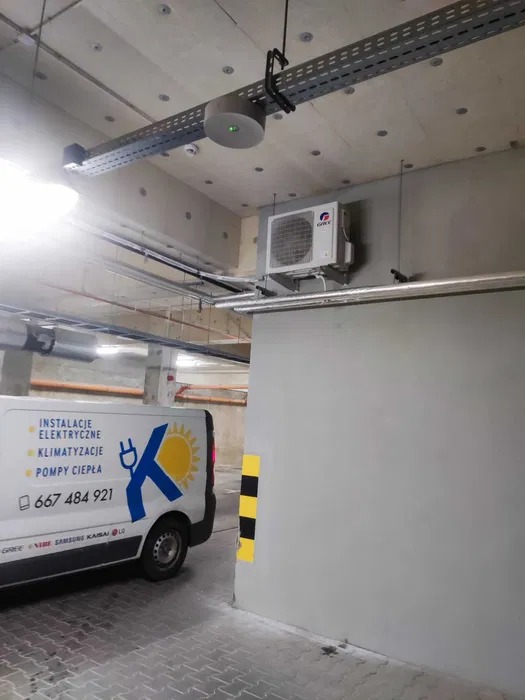 Klimatyzator GREE zamontowany na ścianie w garażu podziemnym, widoczny fragment firmowego samochodu z informacjami o instalacjach elektrycznych, klimatyzacji i pompach ciepła.