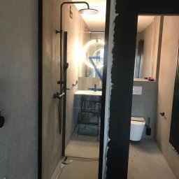 SPLENDIZE SPÓŁKA Z OGRANICZONĄ ODPOWIEDZIALNOŚCIĄ - Nowoczesna kabina prysznicowa typu walk-in z czarnymi profilami, widok z zewnątrz, szara betonowa ściana, częściowo wykończona ściana obok kabiny, widoczny fragment toalety i umywalki w tle.