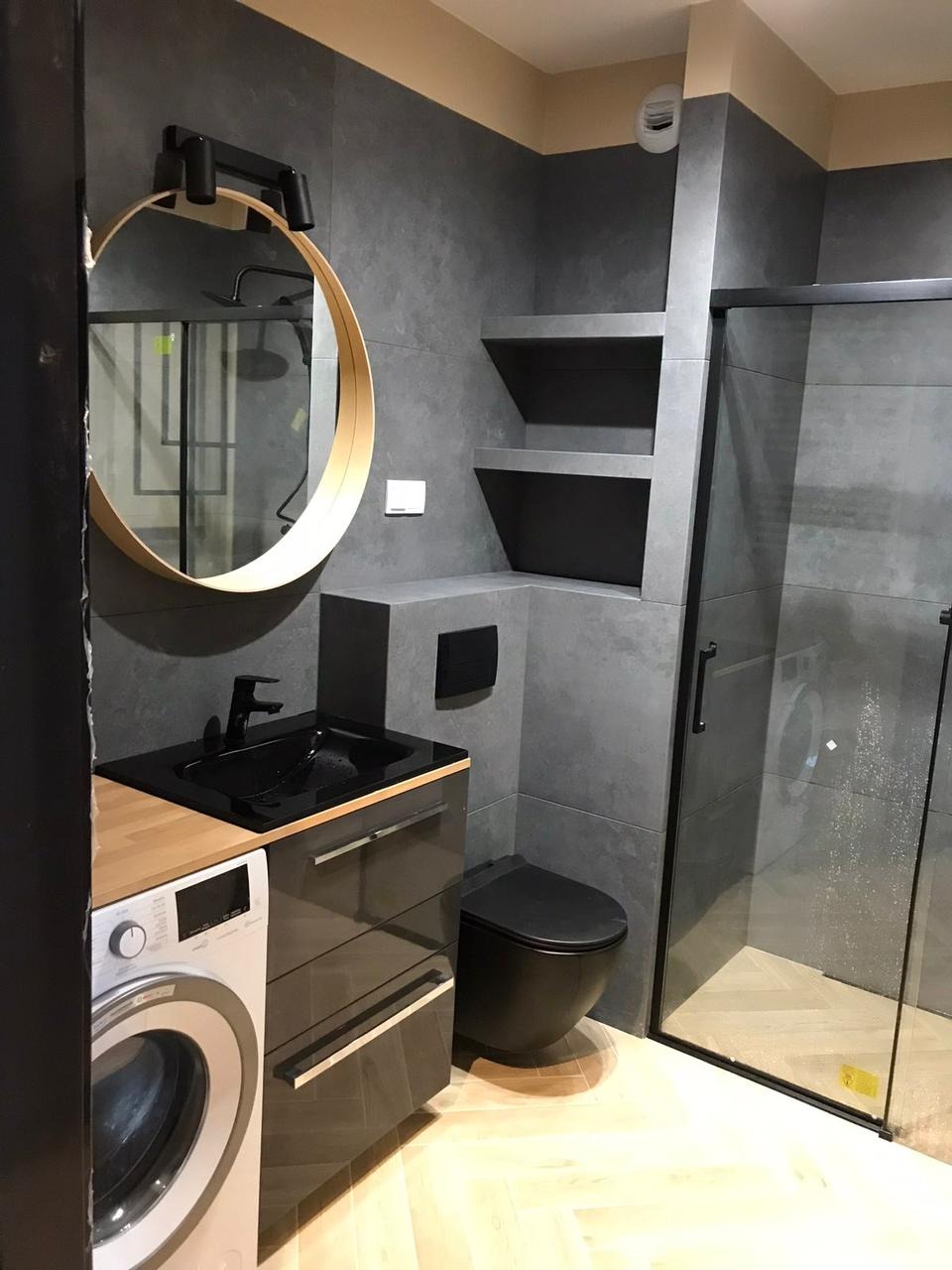 Nowoczesna łazienka z szarymi płytkami, okrągłym lustrem z podświetleniem, czarną armaturą, toaletą i umywalką, pralką obok szafki umywalkowej oraz kabiną prysznicową z czarnymi ramami.