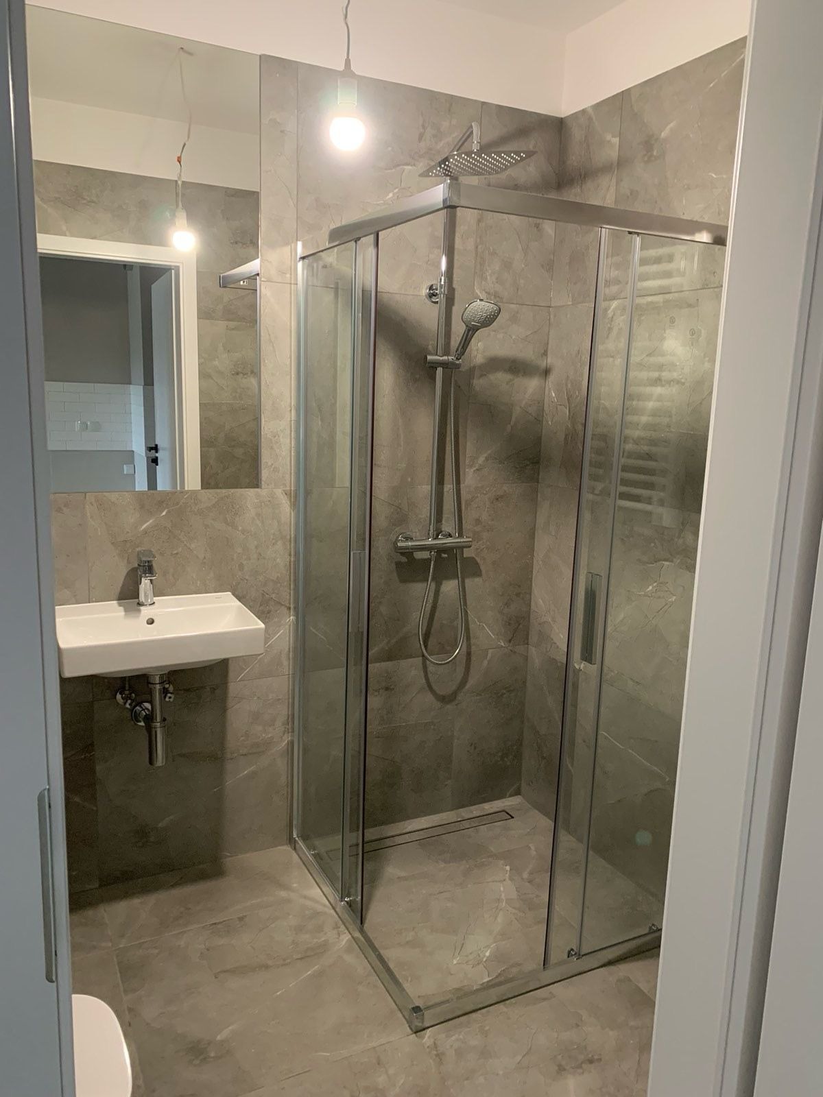 Nowoczesna łazienka w odcieniach szarości z kabiną prysznicową typu walk-in, wiszącą umywalką i dużym lustrem, oświetlona wiszącą żarówką.