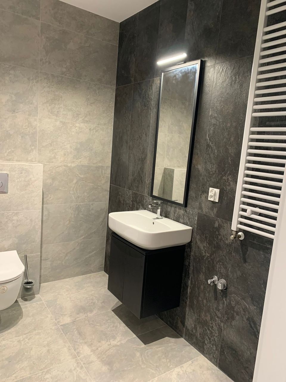 Nowoczesna łazienka w Katowicach z szarymi i grafitowymi płytkami, wisząca szafka z białą umywalką i grzejnik drabinkowy, minimalistyczny styl.