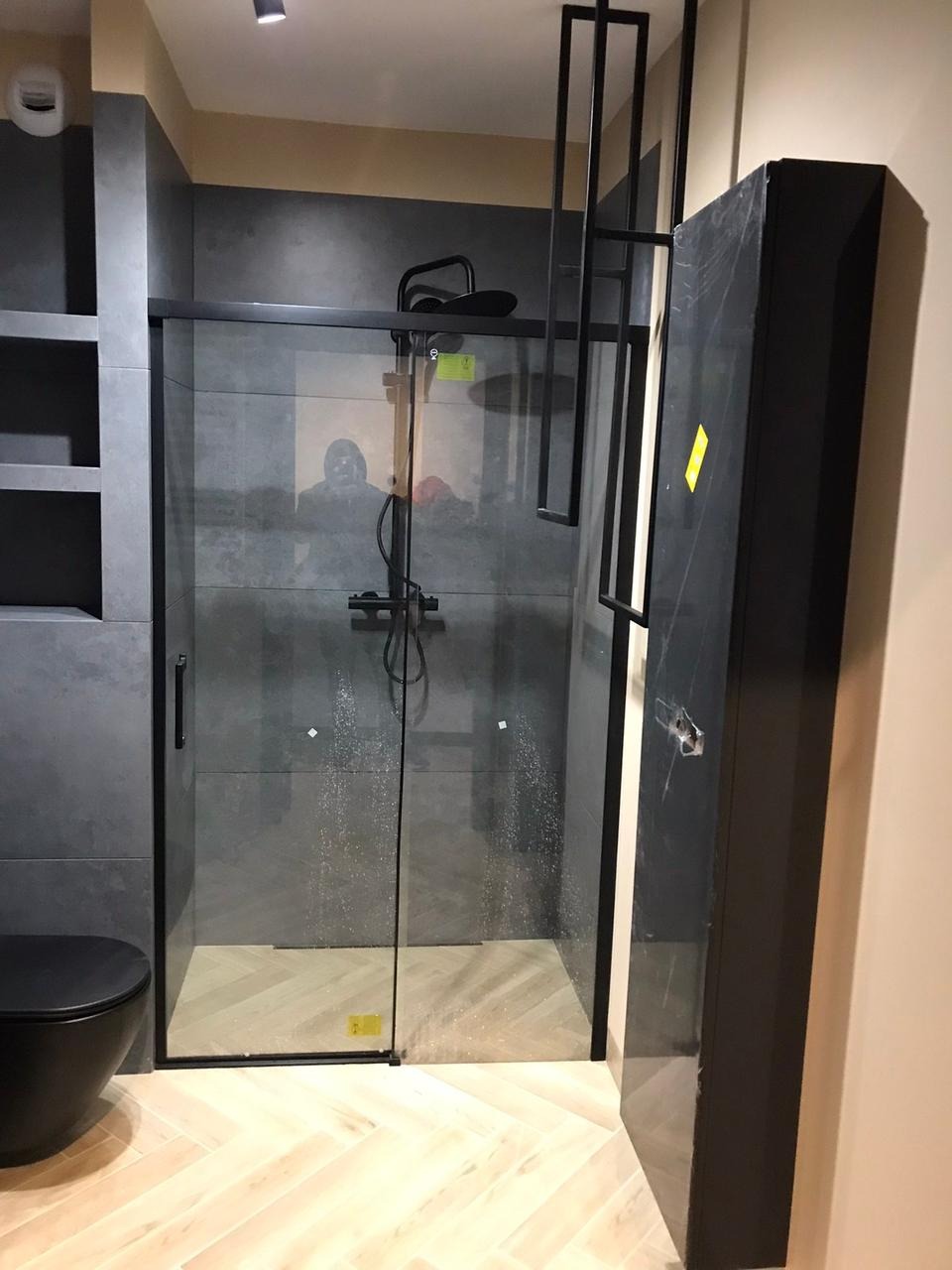 Nowoczesna kabina prysznicowa z czarnymi profilami i deszczownicą, szare płytki na ścianach, podłoga wyłożona parkietem w jodełkę, czarna szafka obok kabiny.