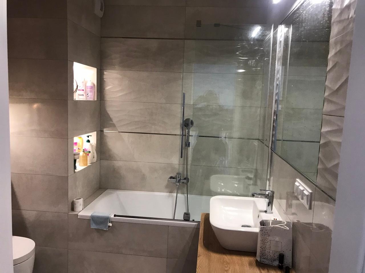 Nowoczesna łazienka z wanną, szklanym parawanem nawannowym, umywalką nablatową na drewnianym blacie i wnękami w ścianie z kosmetykami, wykończona szarymi płytkami o delikatnej fakturze.