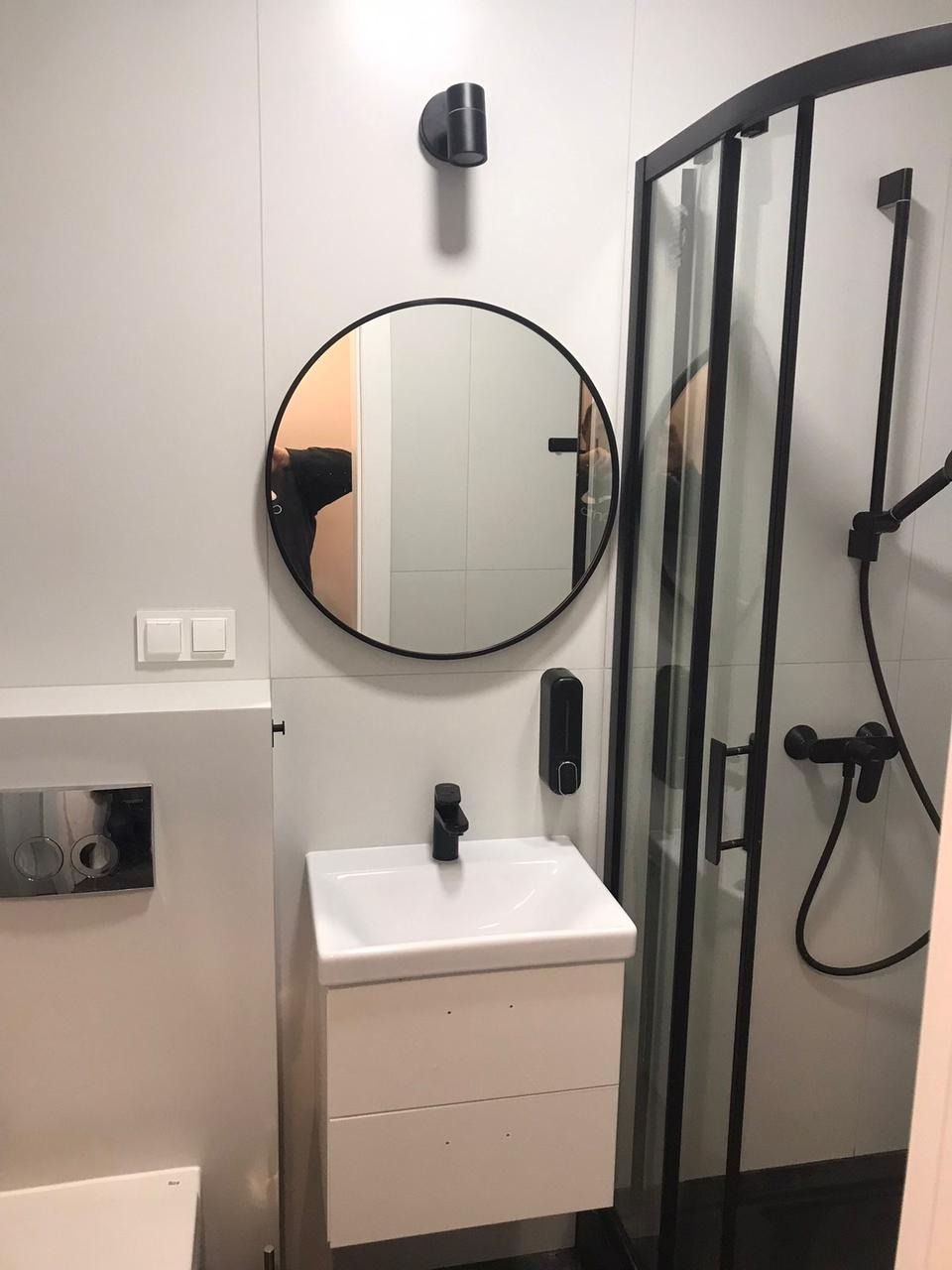 Nowoczesna łazienka z białą armaturą, czarną baterią umywalkową, okrągłym lustrem w czarnej ramie i kabiną prysznicową z czarnymi profilami.