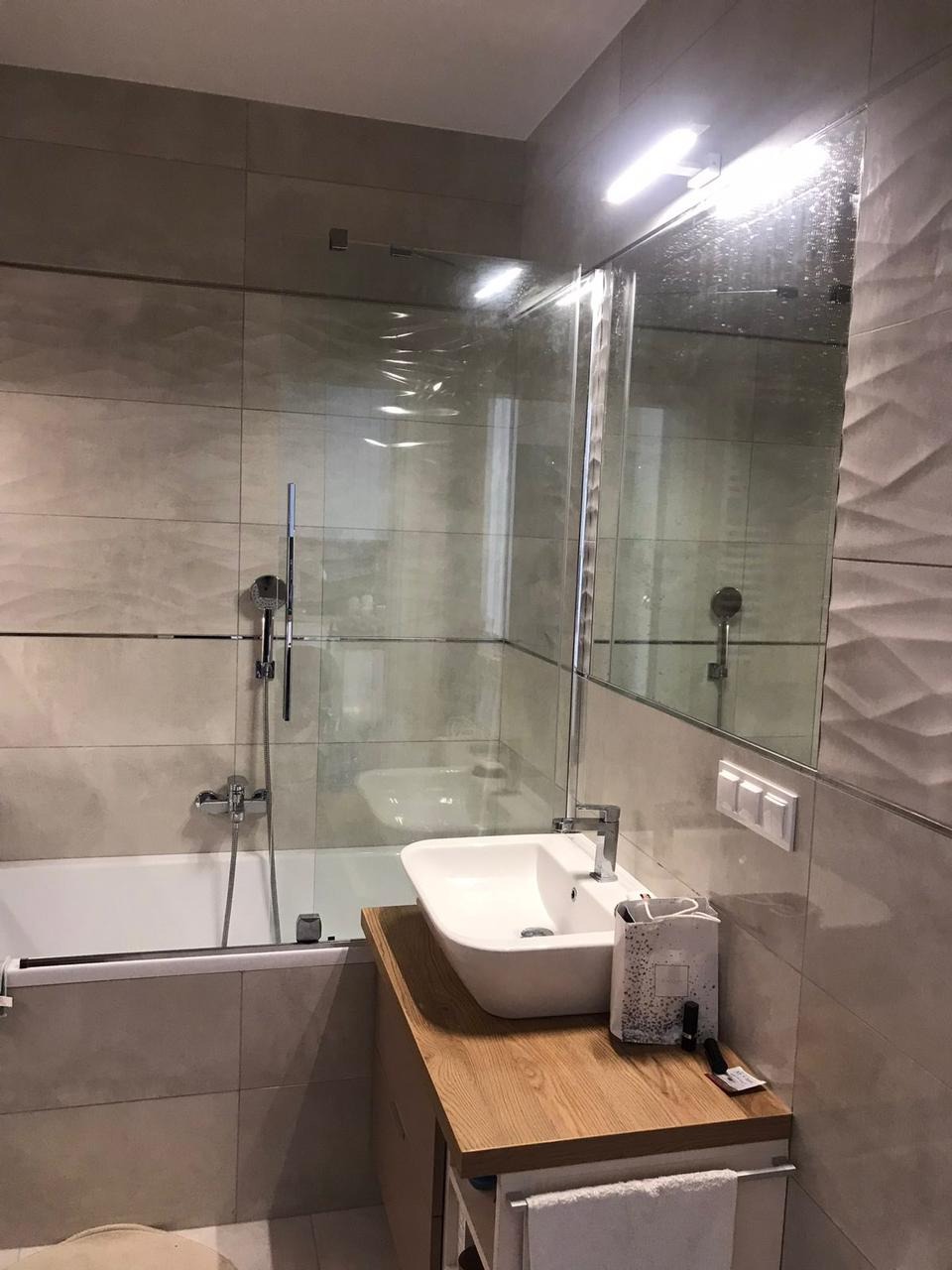 Nowoczesna łazienka z wanną za szklanym panelem, umywalką nablatową na drewnianym blacie i beżowymi płytkami o falistej strukturze, oświetlona lampą LED.