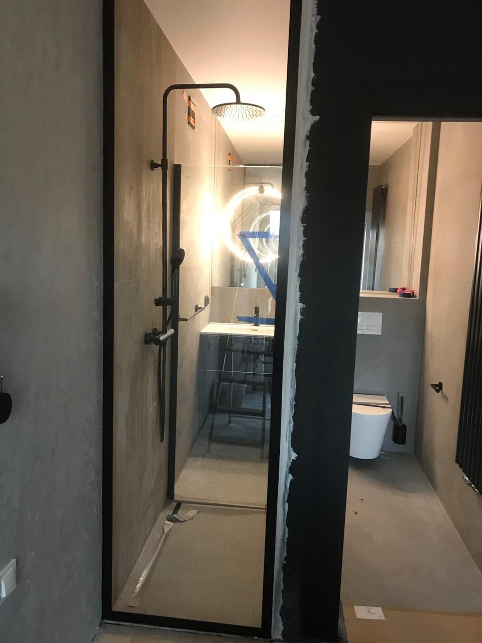 Nowoczesna kabina prysznicowa typu walk-in z czarnymi profilami, widok z zewnątrz, szara betonowa ściana, częściowo wykończona ściana obok kabiny, widoczny fragment toalety i umywalki w tle.