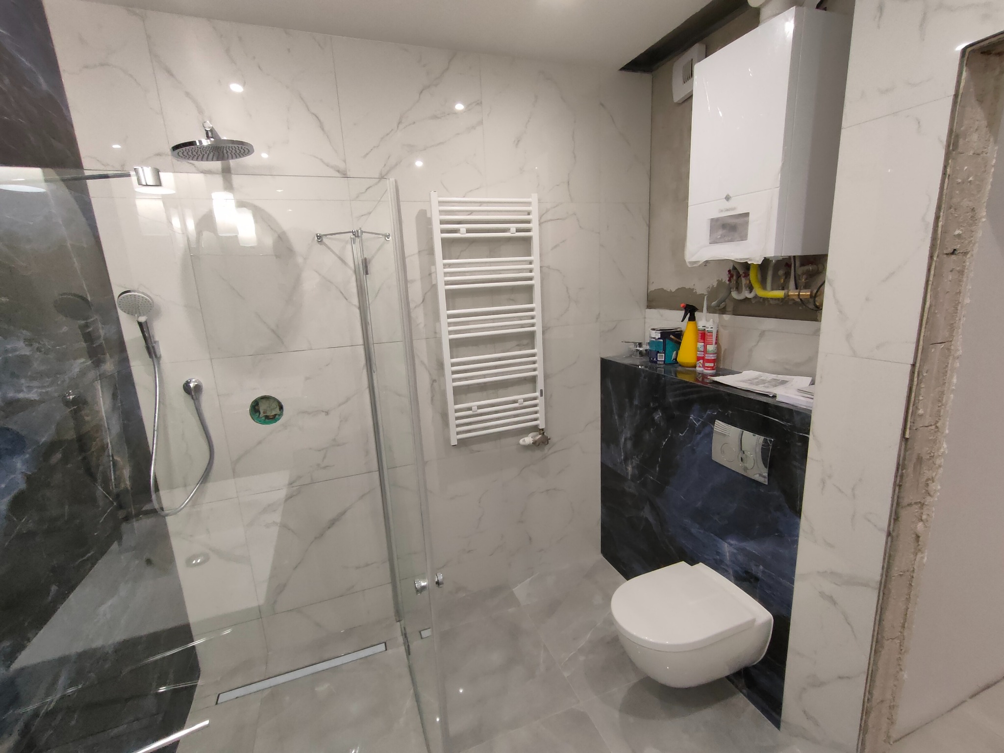 Nowoczesna łazienka z kabiną prysznicową typu walk-in, grzejnikiem drabinkowym, podwieszaną toaletą na tle marmurowych płytek, z widocznym piecem gazowym i akcesoriami instalacyjnymi w trakcie...