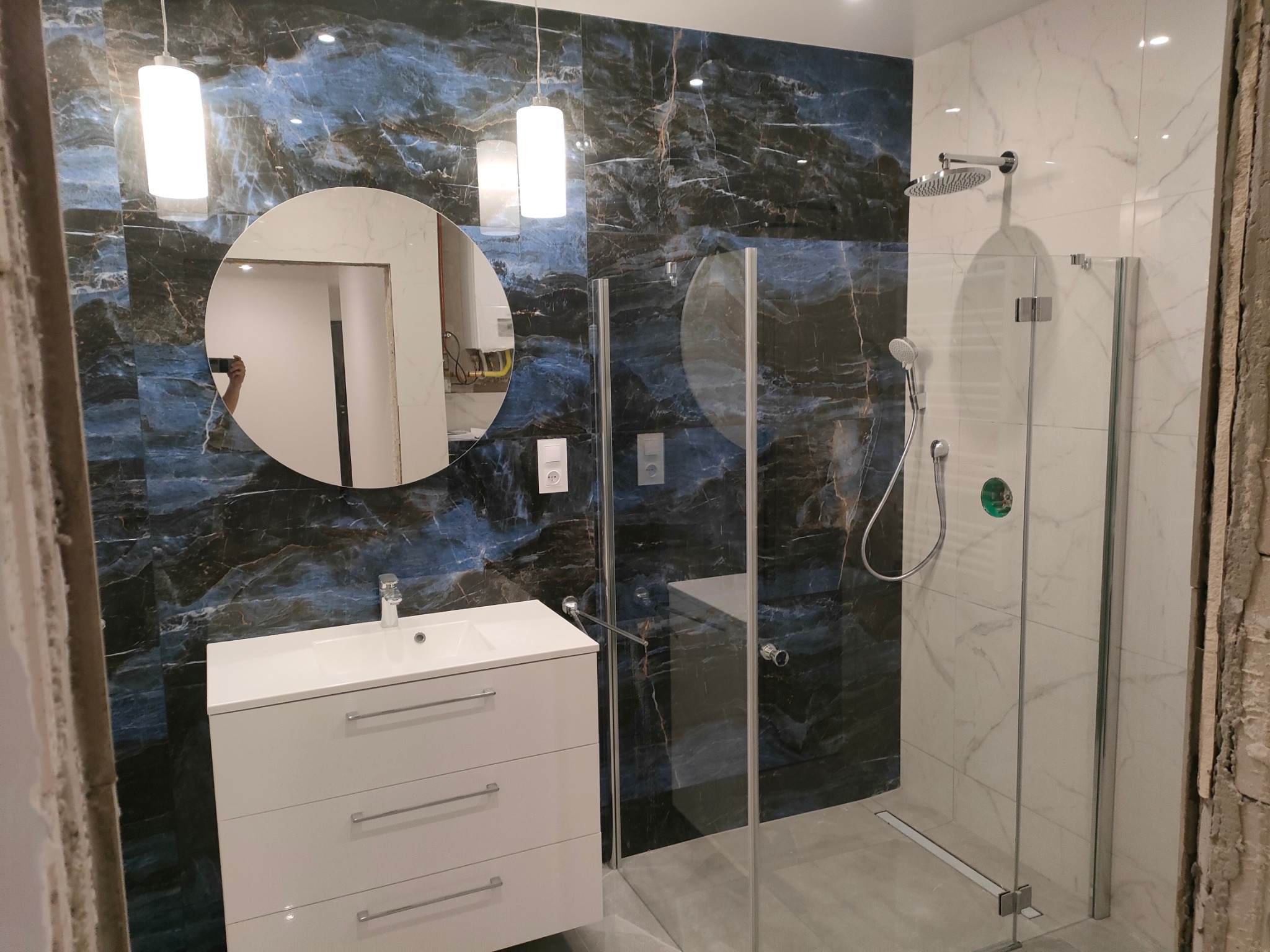 Nowoczesna łazienka z kabiną prysznicową typu walk-in, umywalką z szafką w kolorze białym i okrągłym lustrem, wykończona płytkami w kolorze granatowo-białym imitującymi marmur oraz białymi płytkami...