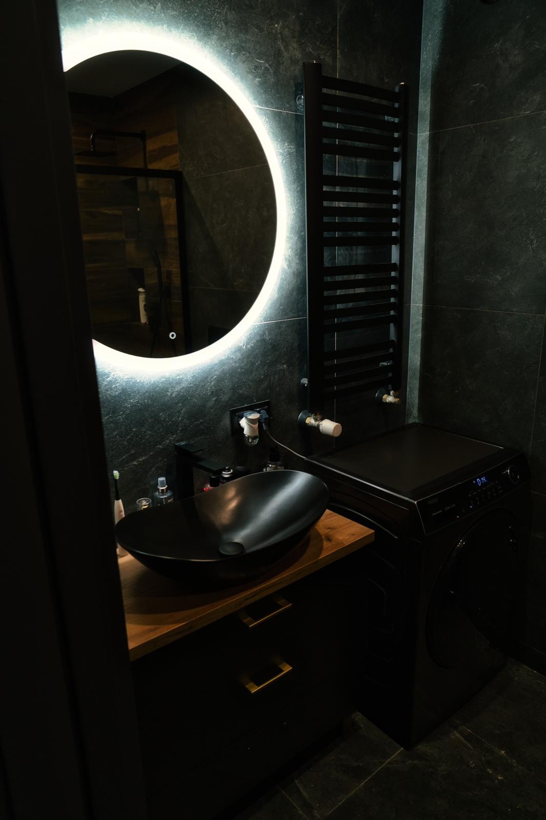 Nowoczesna łazienka z czarnymi płytkami, okrągłym lustrem z podświetleniem LED, czarną umywalką nablatową na drewnianym blacie, czarnym grzejnikiem drabinkowym i pralką w kolorze czarnym.