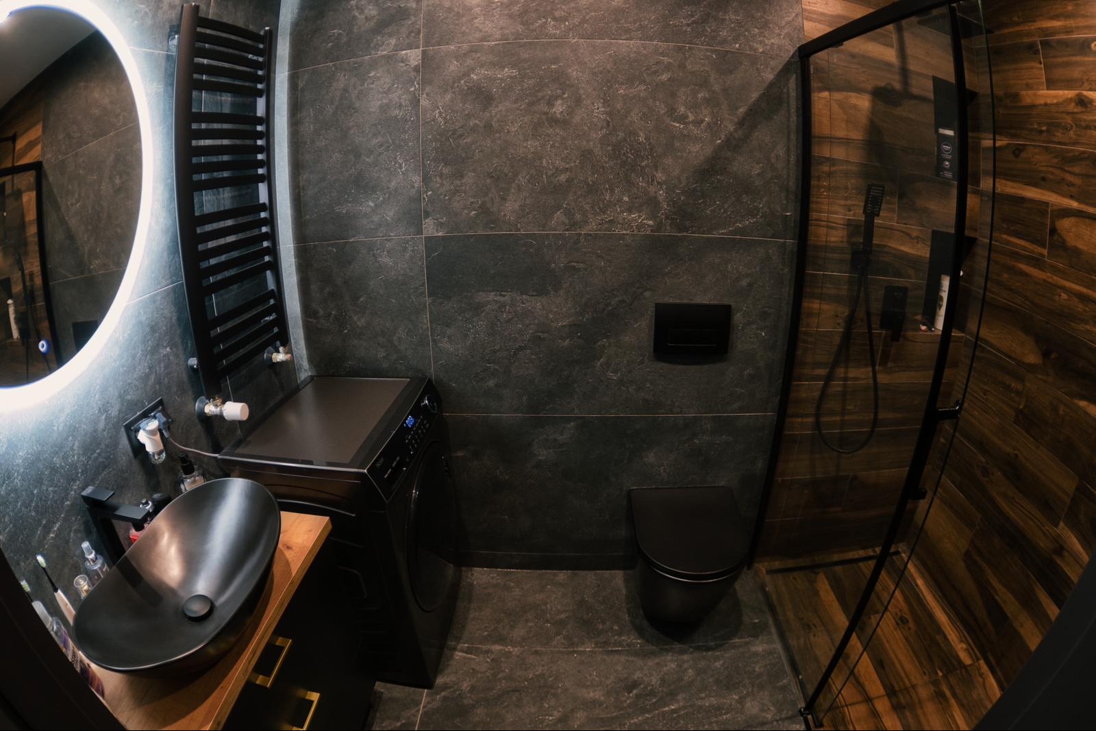 Nowoczesna łazienka z czarną armaturą, umywalką nablatową na drewnianym blacie, grzejnikiem drabinkowym, pralką, toaletą i kabiną prysznicową z drewnopodobnymi płytkami.