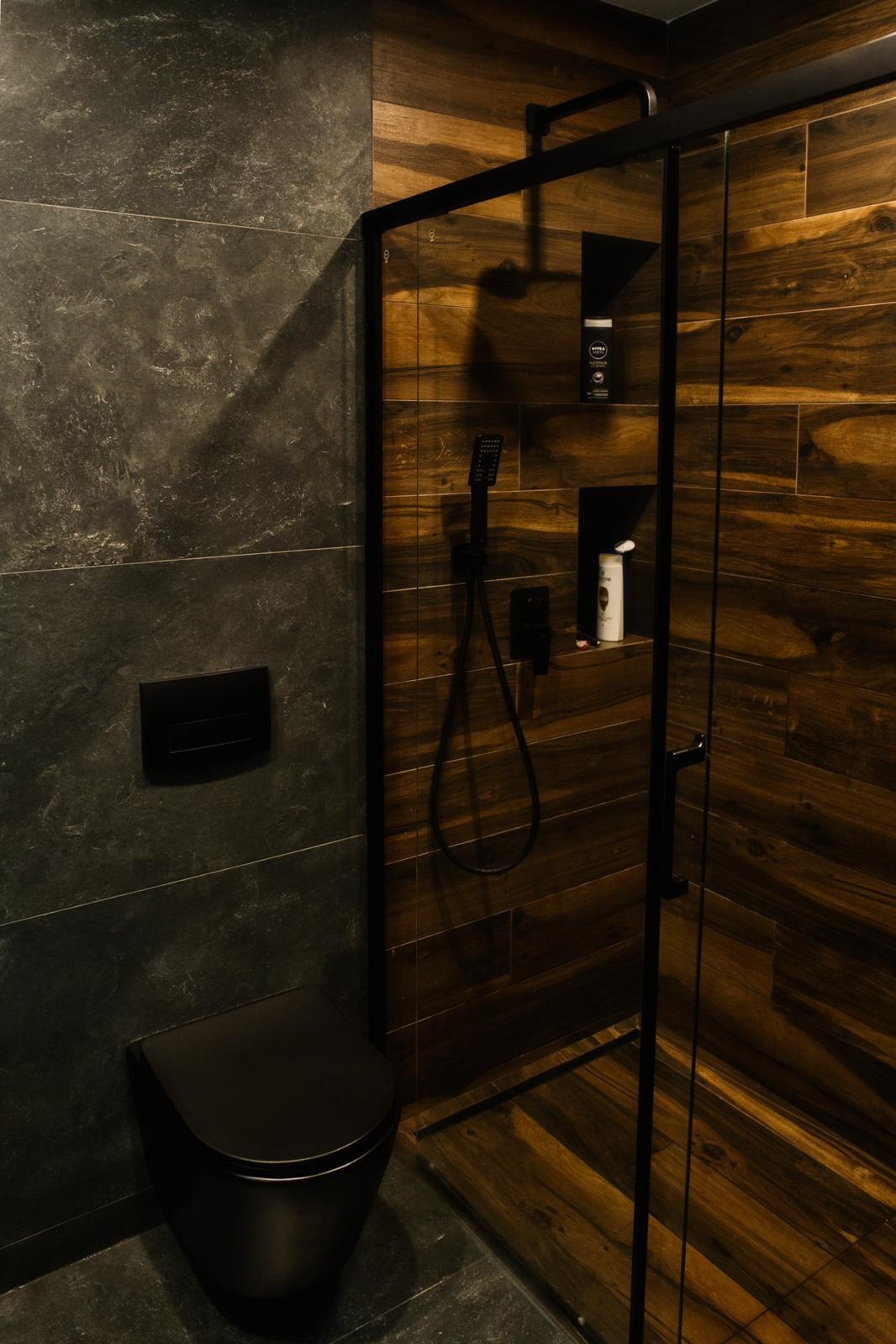 Nowoczesna łazienka z czarną armaturą: kabina prysznicowa z czarnymi ramami, deszczownica, toaleta w kolorze czarnym, ściany wyłożone płytkami imitującymi drewno i szarym kamieniem.