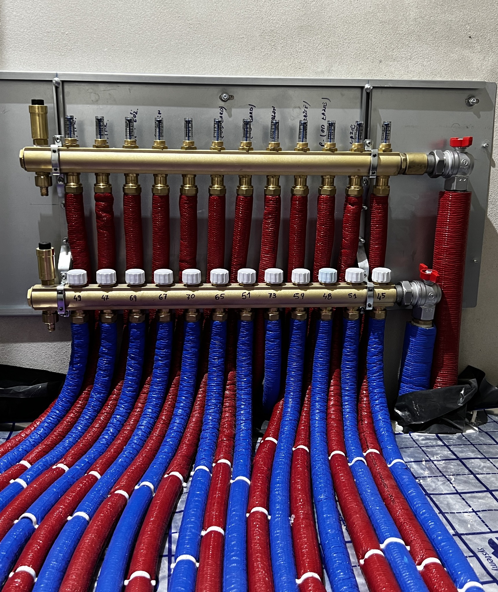 Rozdzielacz ogrzewania podłogowego z czerwonymi i niebieskimi rurami, widoczne numery na belce rozdzielacza oraz przepływomierze z odczytami, zamontowany na szarej ścianie.