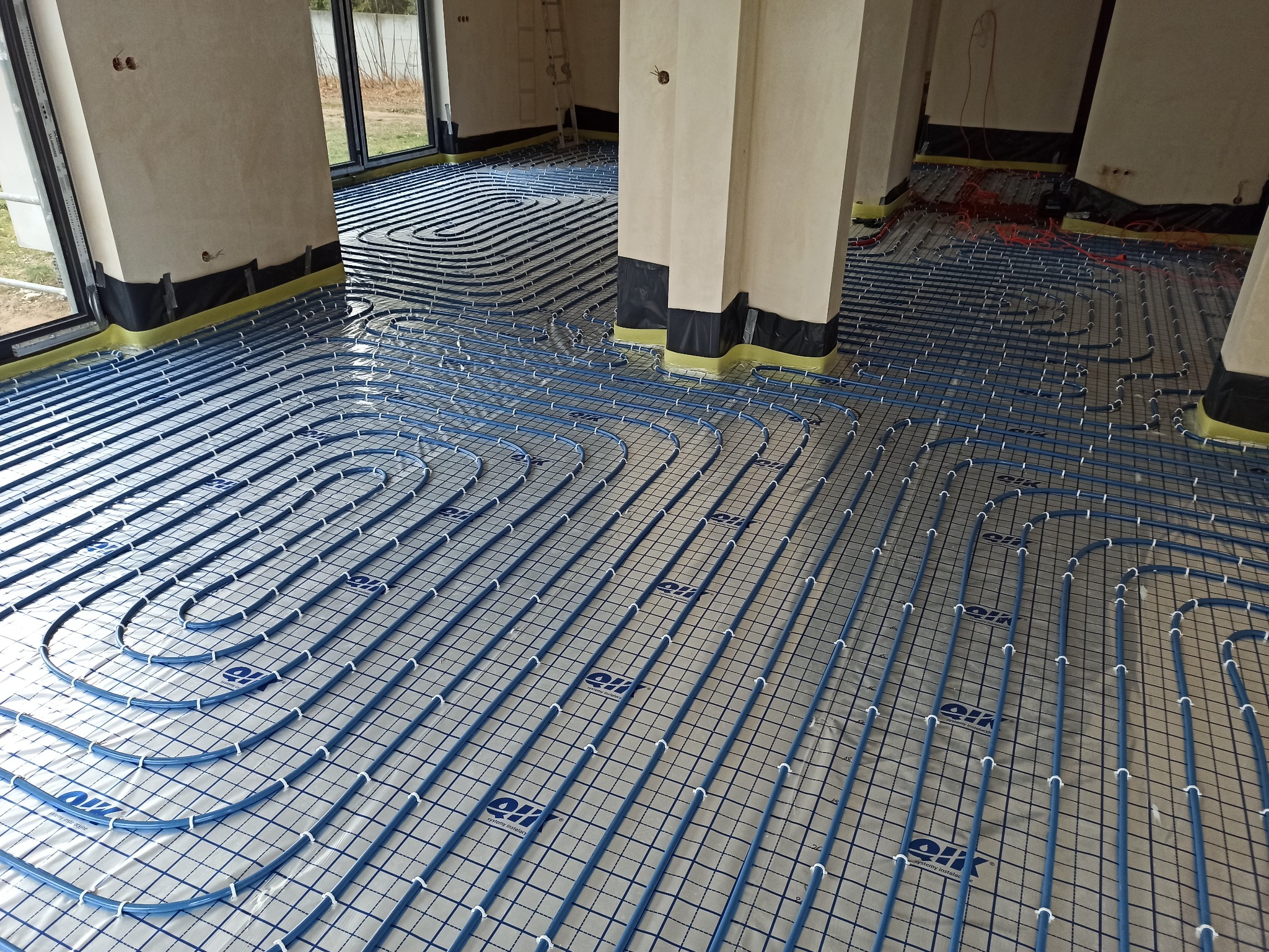 Niebieskie rury ogrzewania podłogowego ułożone na folii z nadrukowaną siatką, widoczne fragmenty ścian i kolumn zabezpieczonych folią ochronną.