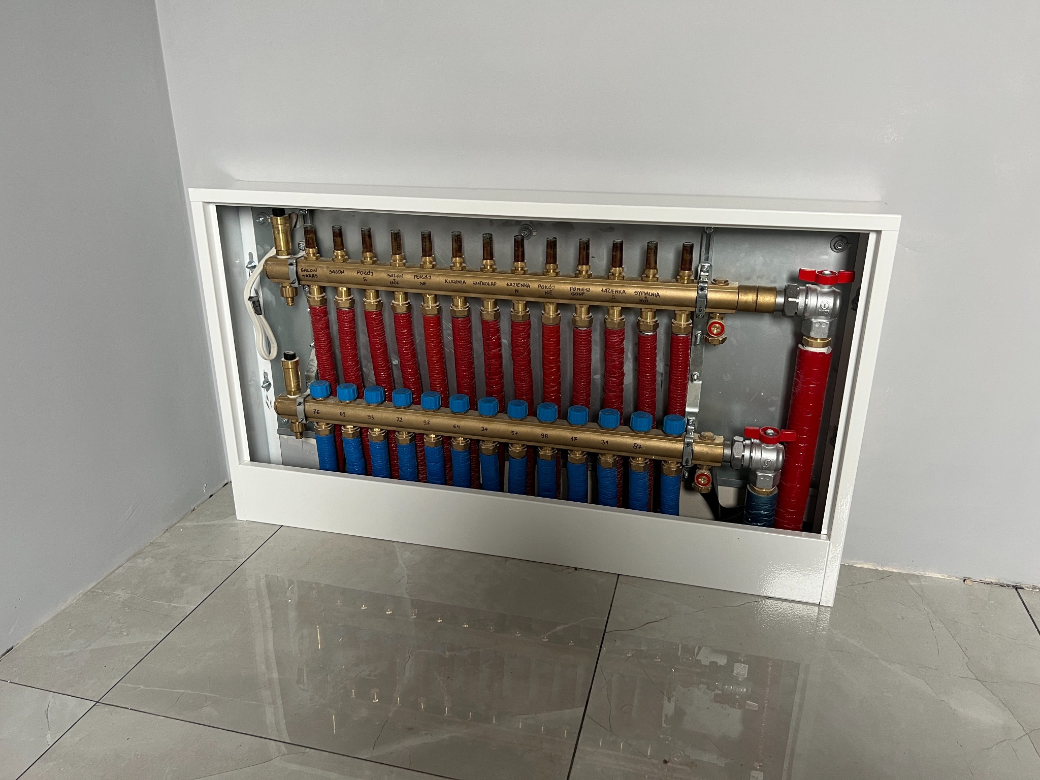 Rozdzielacz ogrzewania podłogowego w białej obudowie, widoczne rury w kolorze czerwonym i niebieskim, z opisami pomieszczeń na górnej listwie, zamontowany na tle szarej ściany i płytek podłogowych.