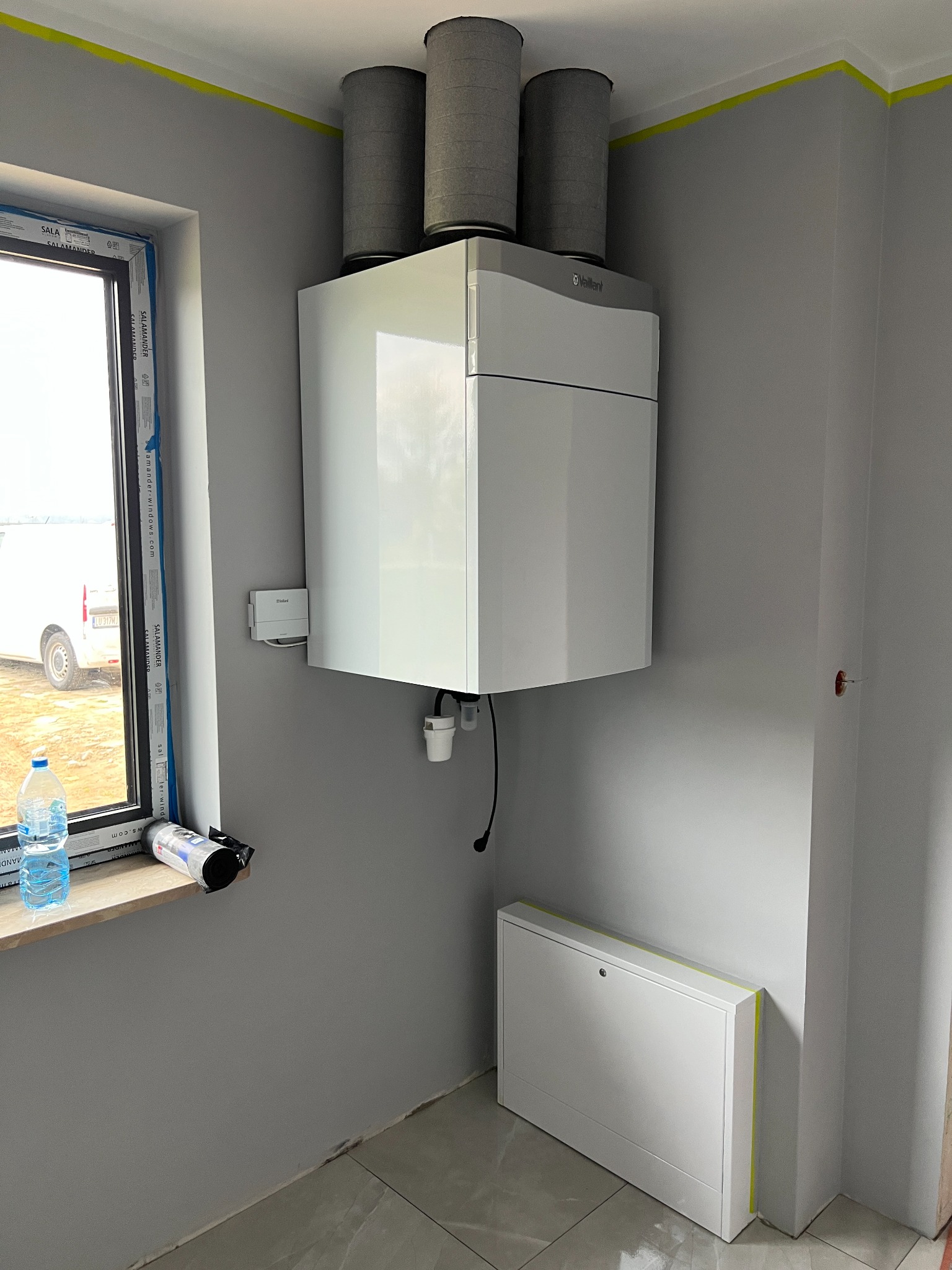 Biała jednostka centralna rekuperacji marki Vaillant z trzema szarymi filtrami cylindrycznymi, zamontowana na szarej ścianie w surowym wnętrzu, z widocznym oknem i butelkami wody na parapecie...