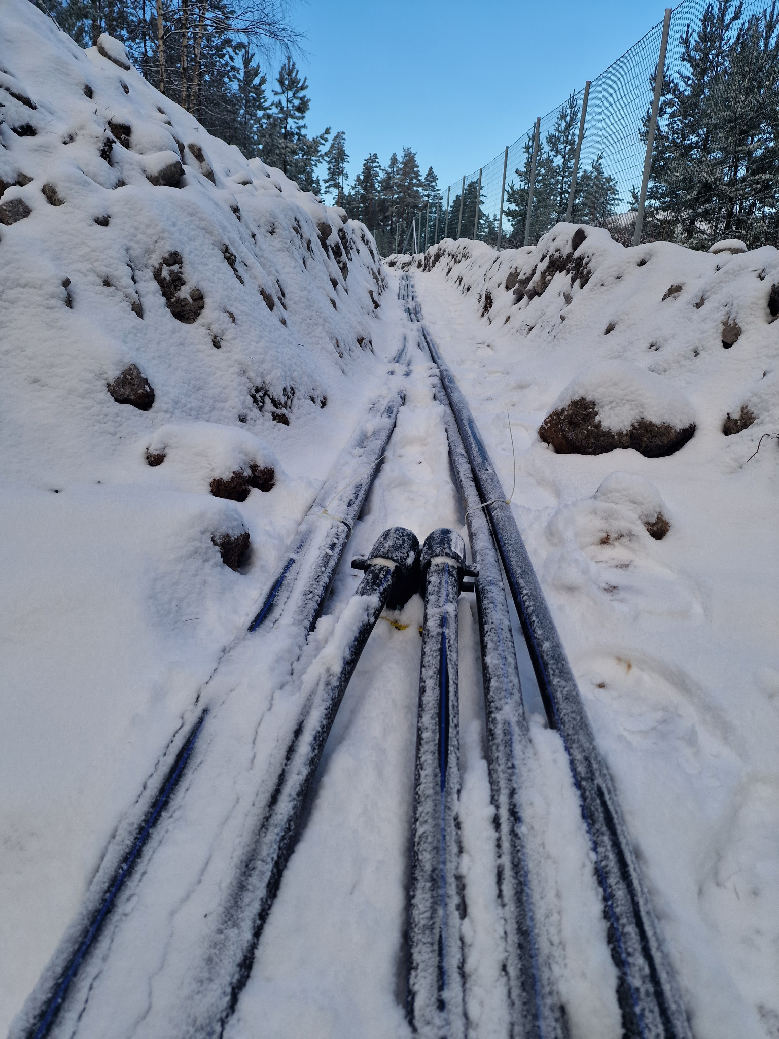 Rury zakopane w śniegu w wykopie, widoczne złącza, w tle siatka ogrodzeniowa i drzewa.