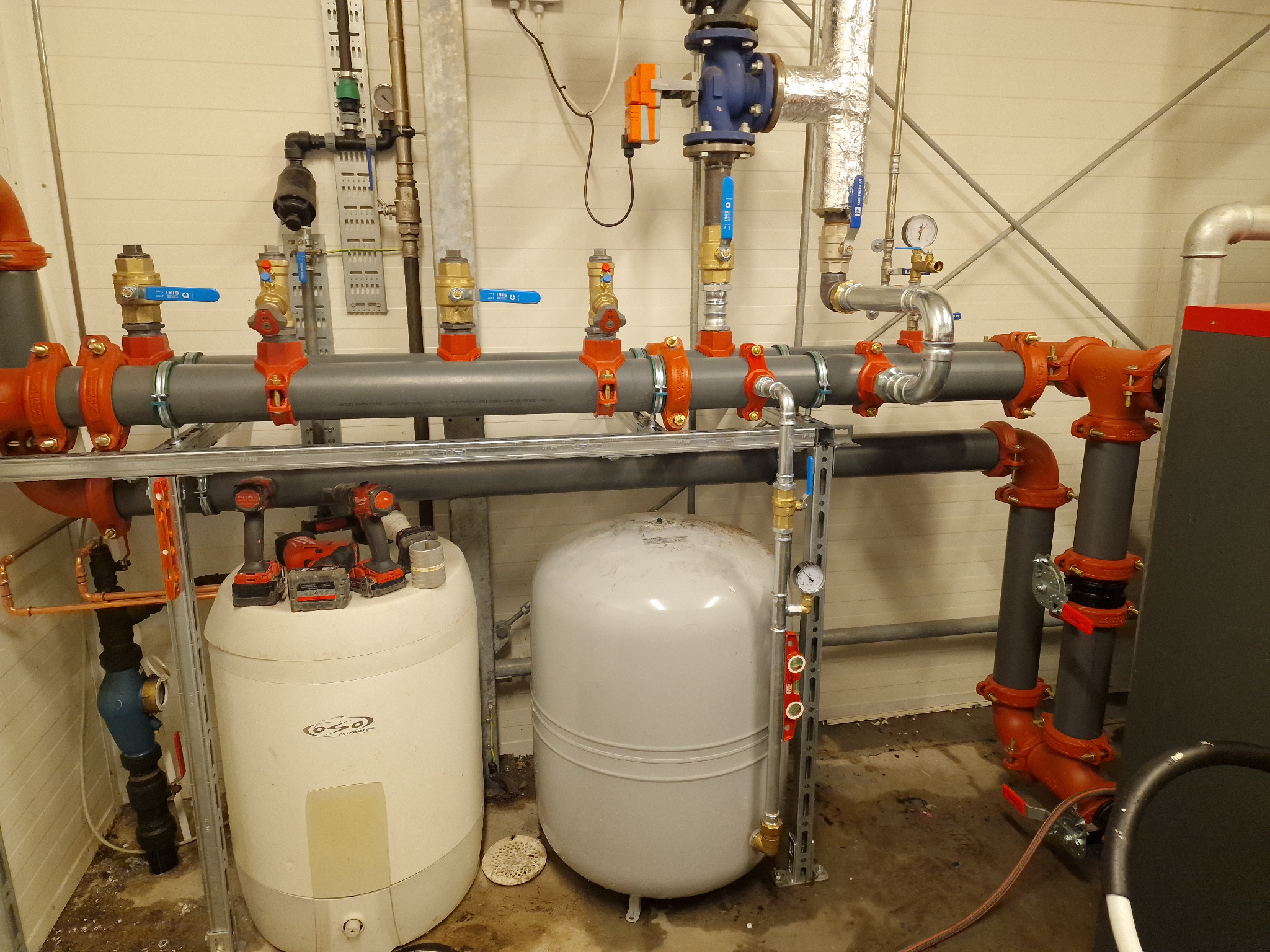 Rozbudowana instalacja hydrauliczna w kotłowni: rury, zawory, zbiorniki wyrównawcze, manometry, oraz narzędzia akumulatorowe na zbiorniku.