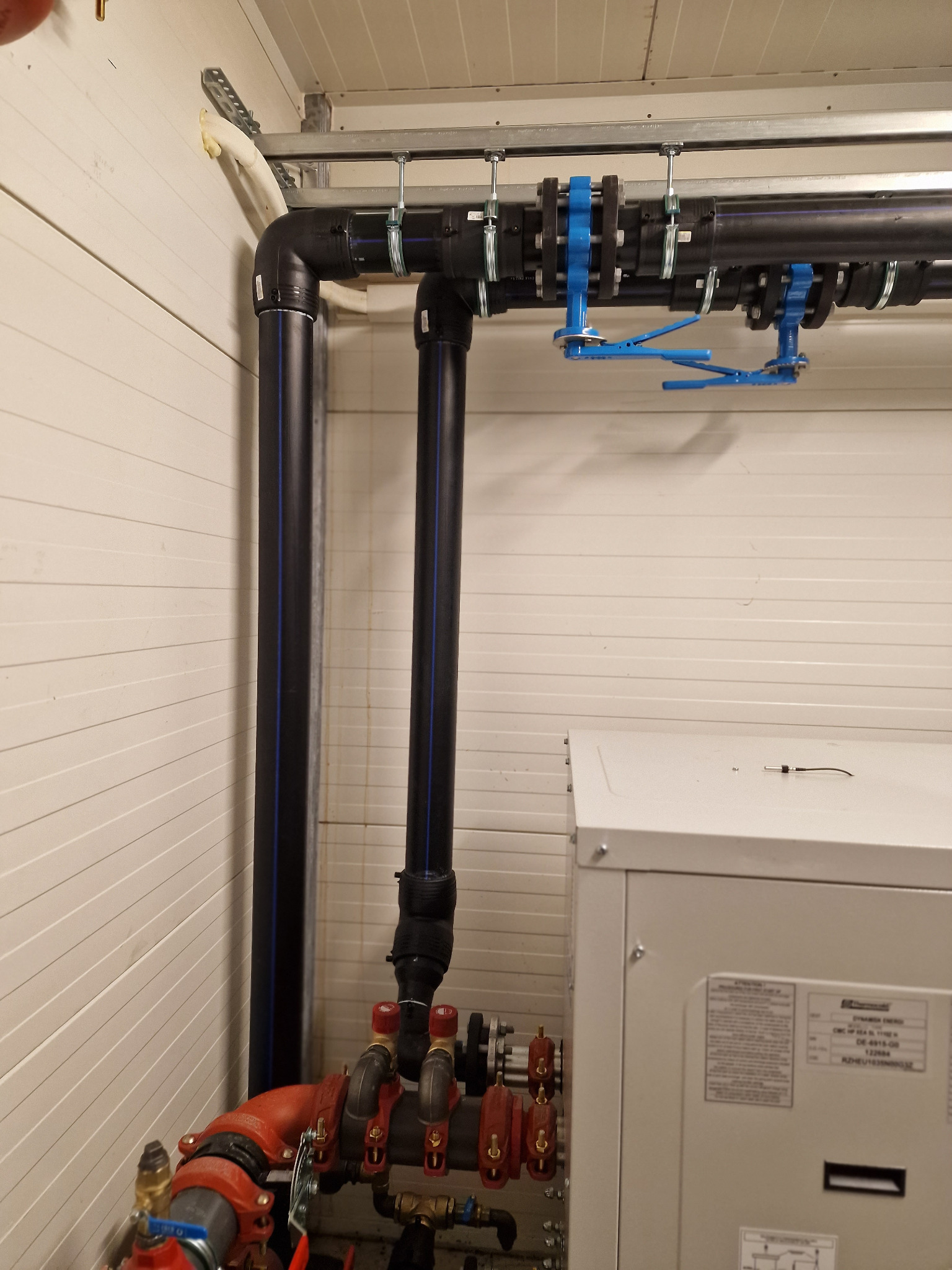 Fragment instalacji z czarnych rur z niebieskim paskiem, mocowanych do sufitu za pomocą metalowych uchwytów i niebieskich zacisków, widoczny fragment czerwonej instalacji i szarej obudowy urządzenia.